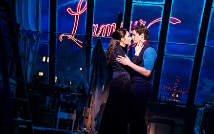 Couple embracing on stage with Moulin Rouge sign in background.