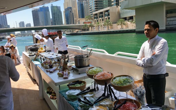 Buffet setup on a yacht with chefs and staff, Dubai Marina skyline in the background.