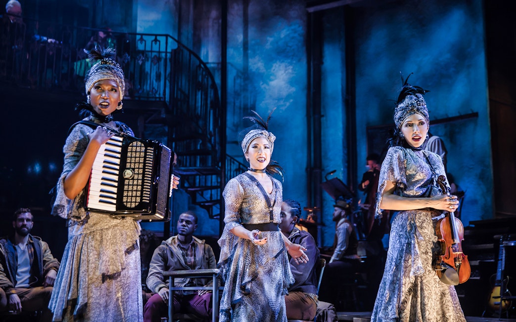 Performers on stage during a Hadestown musical scene with accordion and violin.