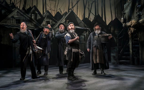 Actors performing a scene from "Fiddler On The Roof" on stage with a village backdrop.