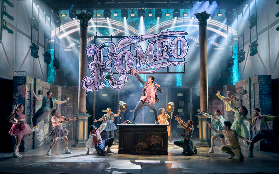 Stage performance of &Juliet with actors dancing under a large "Romeo" sign.