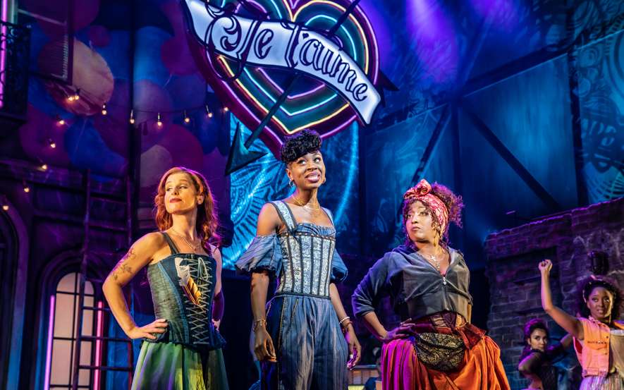Three performers on stage in colorful costumes under a neon "Je t'aime" sign in the &Juliet musical.