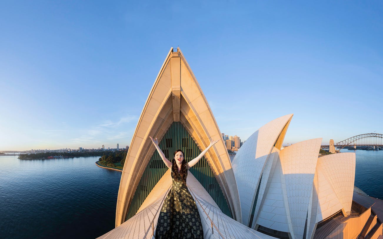 Opera singer on Sydney Opera House roof with city skyline and harbor in background.