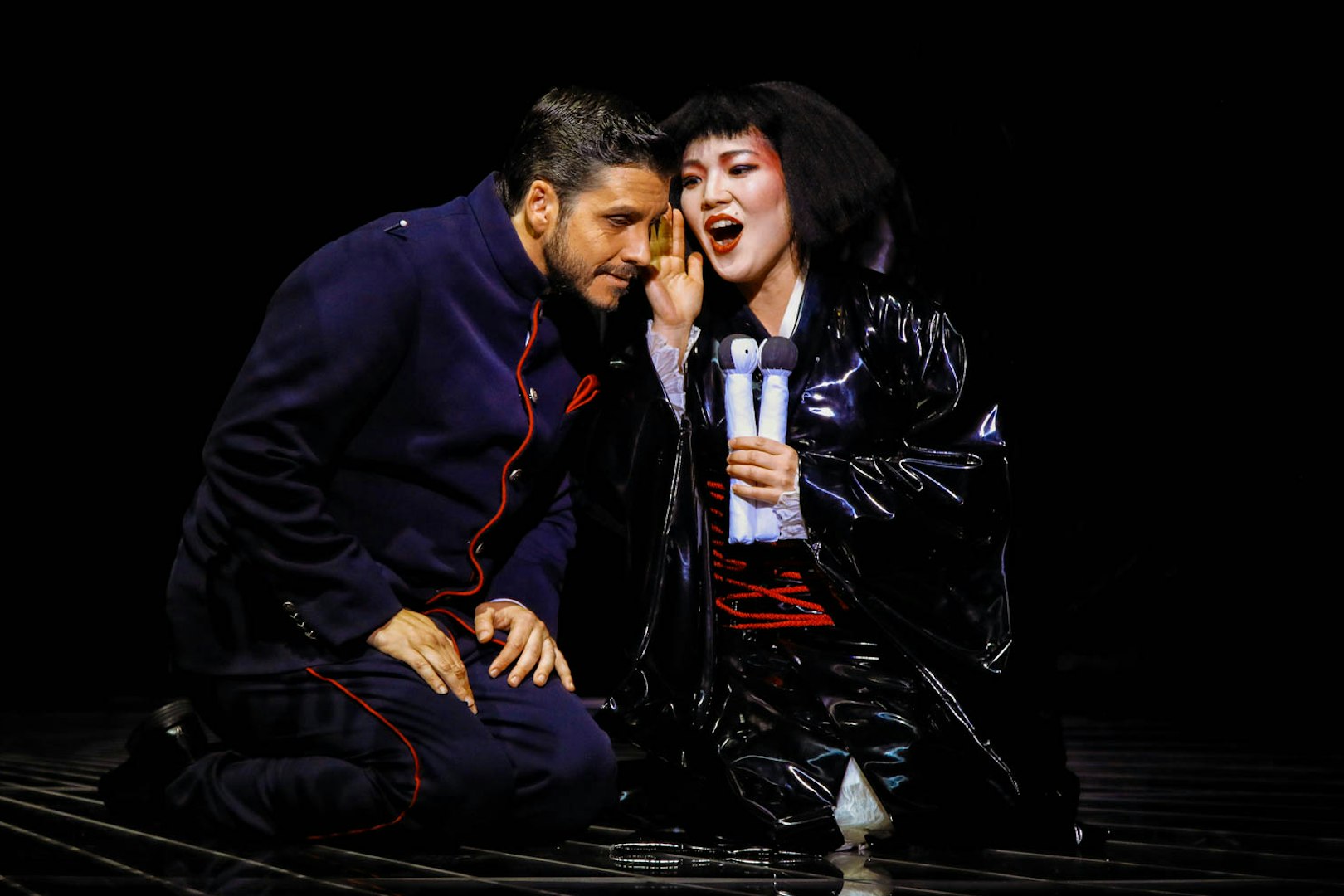 Opera performers in a scene from Madama Butterfly, with one whispering to the other.