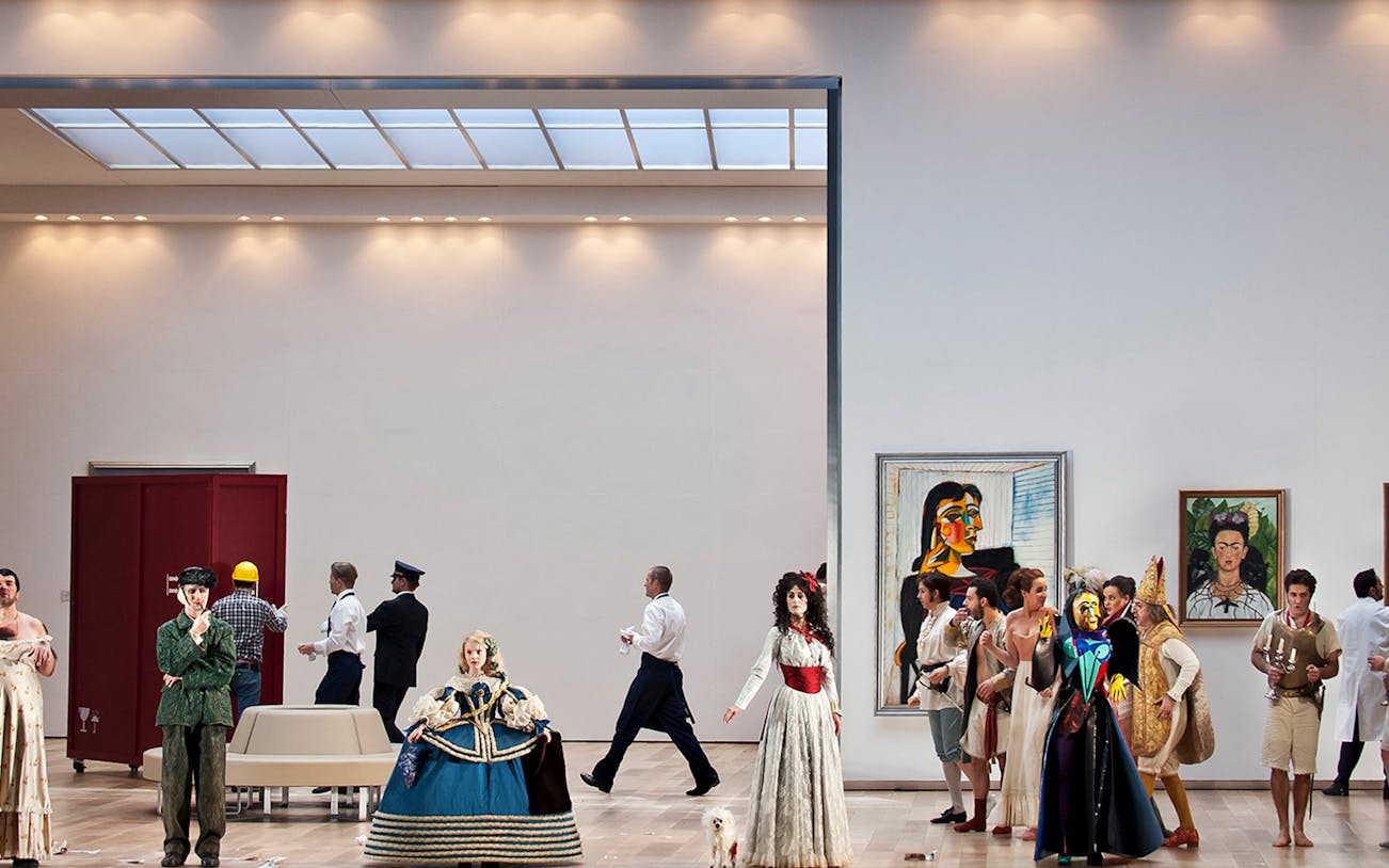 Opera performers in period costumes on stage for "Il viaggio a Reims" with modern art in the background.
