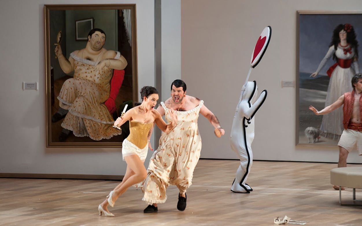 Opera performers in a comedic scene from "Il viaggio a Reims" with art in the background.