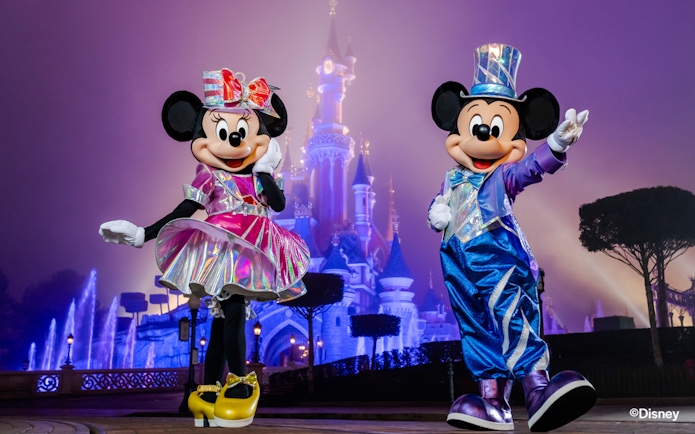 Mickey and Minnie Mouse in festive outfits at Disneyland Paris with Sleeping Beauty Castle in the background.