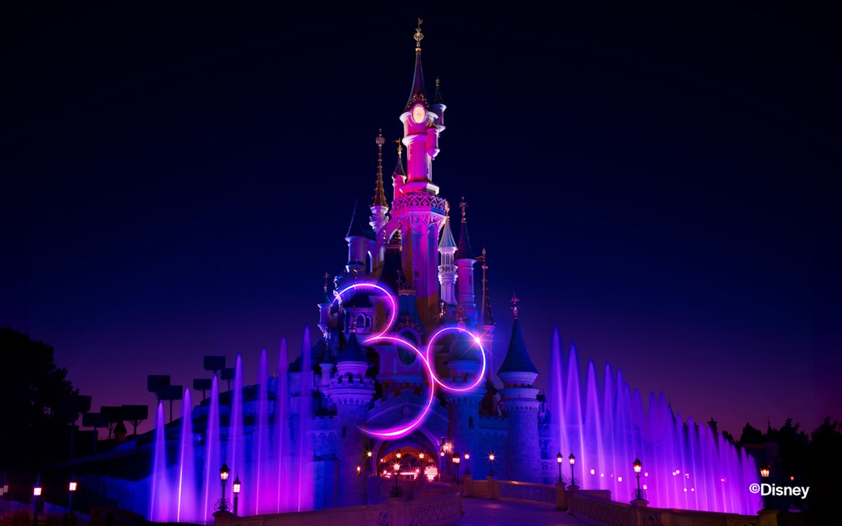 Disneyland Paris castle illuminated at night with colorful fountains.