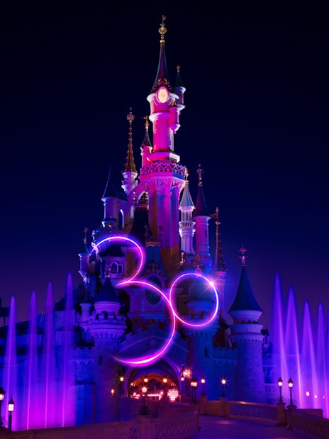 Disneyland Paris castle illuminated at night with colorful fountains.