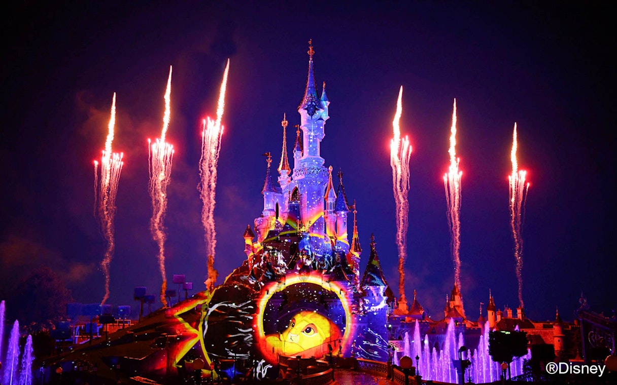 Fireworks over illuminated Disney castle at night, featuring Lion King projection.