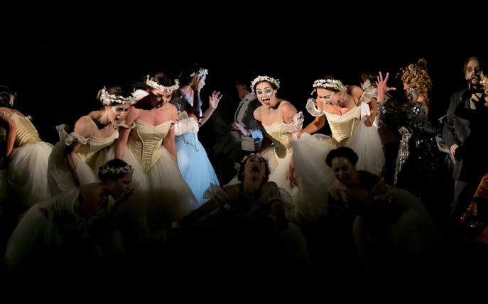 Performers in a dramatic scene from the opera Faust, featuring dancers in white dresses.