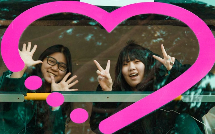 Two people smiling and making peace signs through a bus window with a pink heart design, Rome tour.