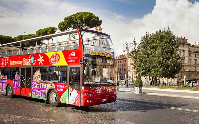 Red double-decker sightseeing bus in Rome near historical buildings and trees.