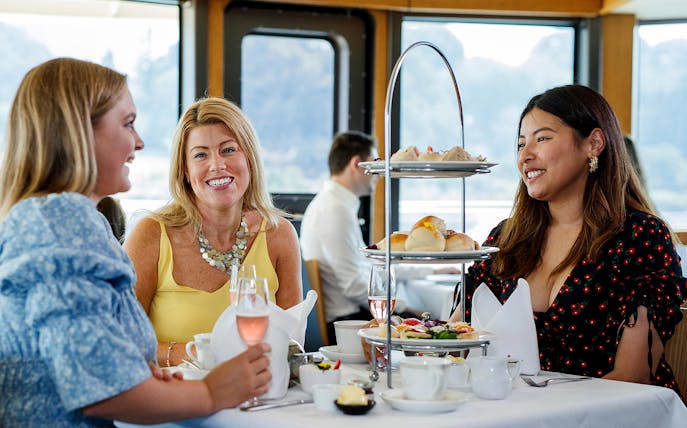 Women enjoying high tea on a cruise with tiered tray of pastries and sandwiches.
