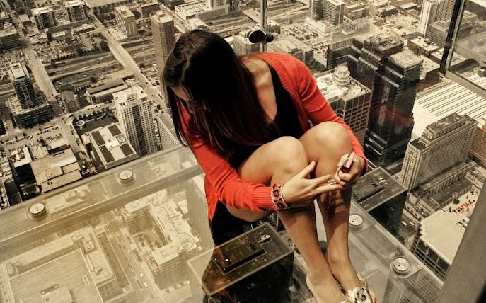 Person sitting on glass ledge at Sky Deck Chicago overlooking cityscape.