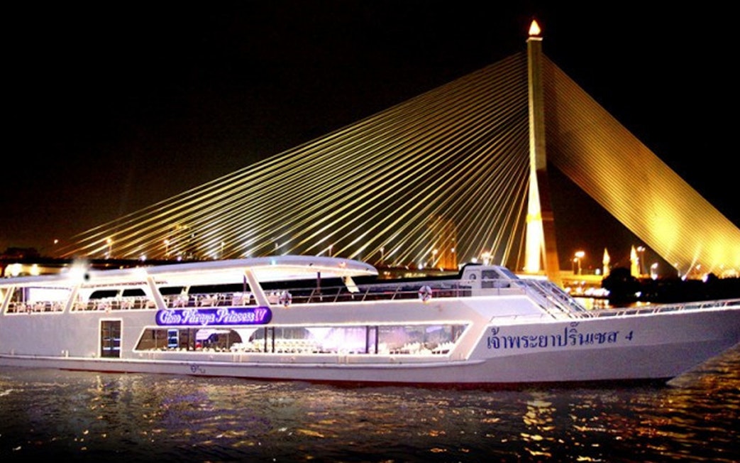 Dinner cruise boat on Chao Phraya River with illuminated bridge in Bangkok.