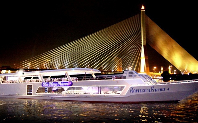 Dinner cruise boat on Chao Phraya River with illuminated bridge in Bangkok.