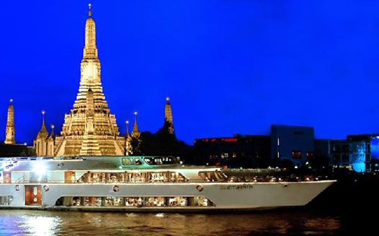 Dinner cruise boat on Chao Phraya River with Wat Arun temple illuminated at night, Bangkok.