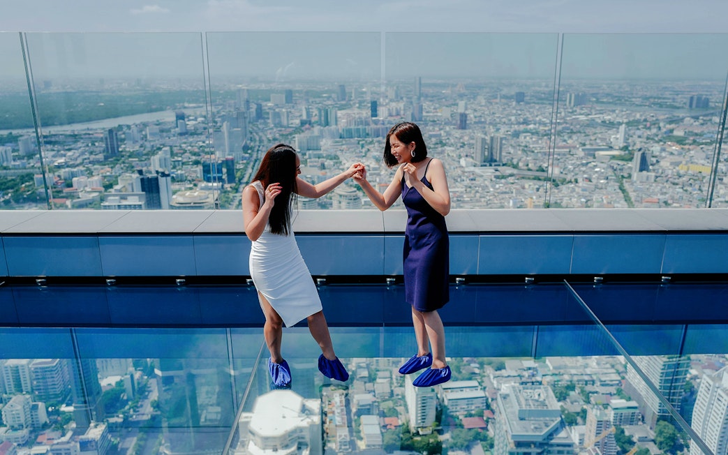 Two people on Mahanakhon SkyWalk glass floor with Bangkok city view.