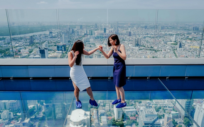 Two people on Mahanakhon SkyWalk glass floor with Bangkok city view.