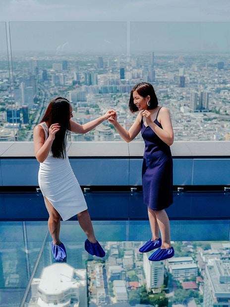 Two people on Mahanakhon SkyWalk glass floor with Bangkok city view.
