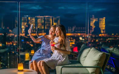 Two people taking a selfie at Mahanakhon SkyWalk, Bangkok, with city lights in the background.