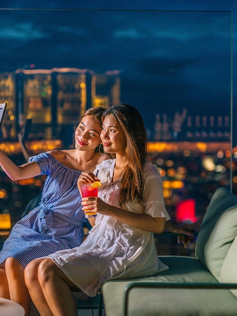 Two people taking a selfie at Mahanakhon SkyWalk, Bangkok, with city lights in the background.