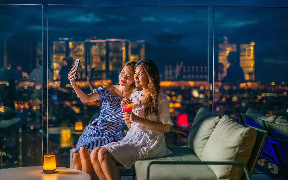 Two people taking a selfie at Mahanakhon SkyWalk, Bangkok, with city lights in the background.