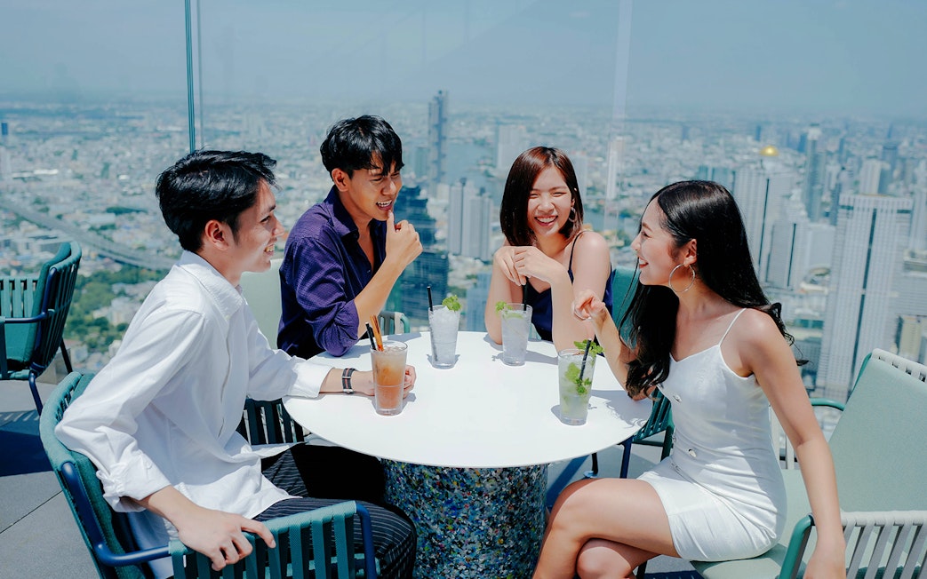 Group enjoying drinks at Mahanakhon SkyWalk with Bangkok skyline in the background.