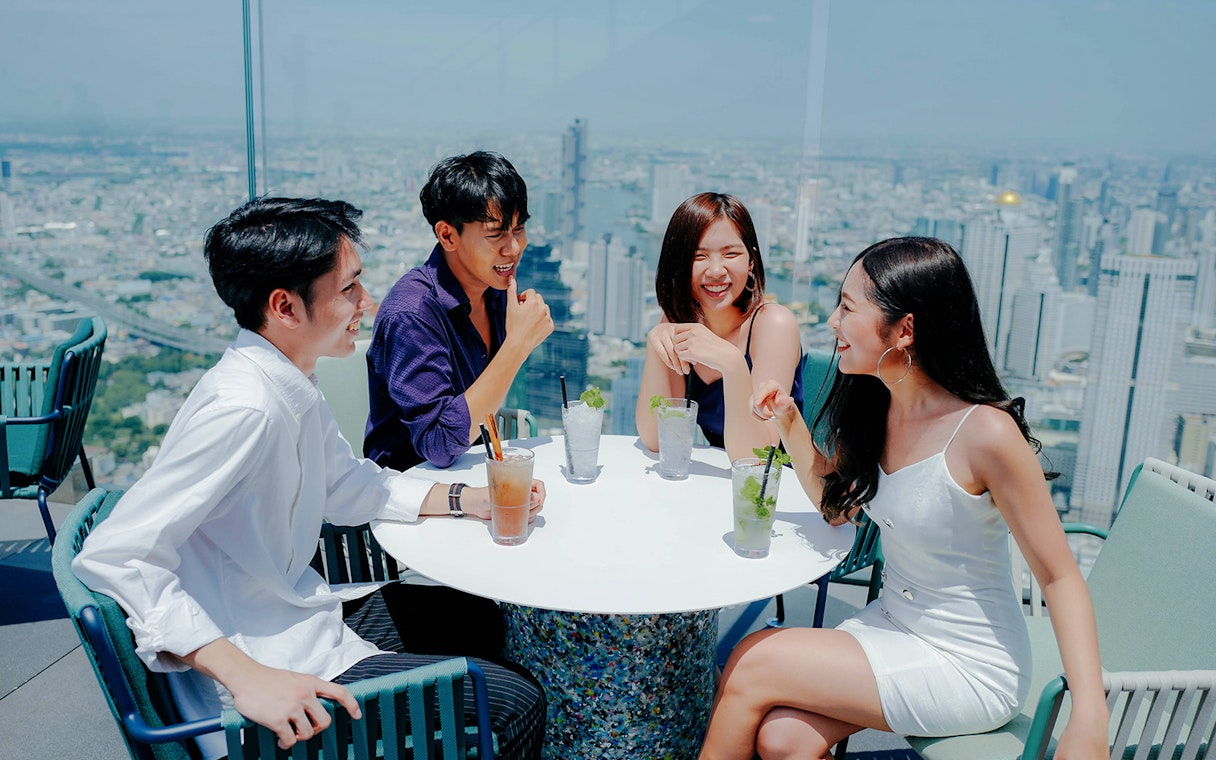 Group enjoying drinks at Mahanakhon SkyWalk with Bangkok skyline in the background.