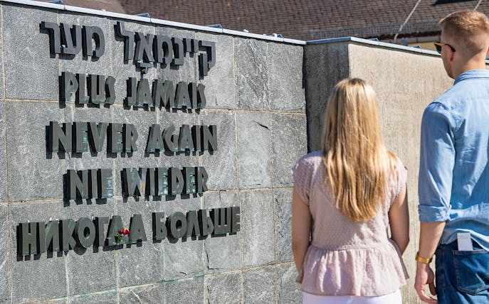 Visitors at Dachau Concentration Camp memorial wall with "Never Again" inscription.