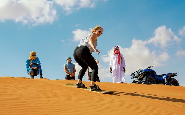 Person sandboarding on red dunes with onlookers and ATV in desert.