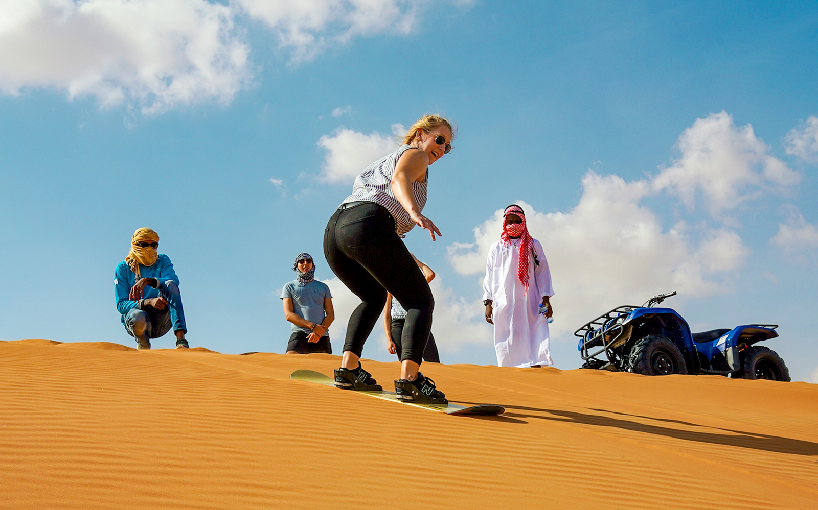 Sandboarding on desert dunes with quad bike and onlookers nearby.