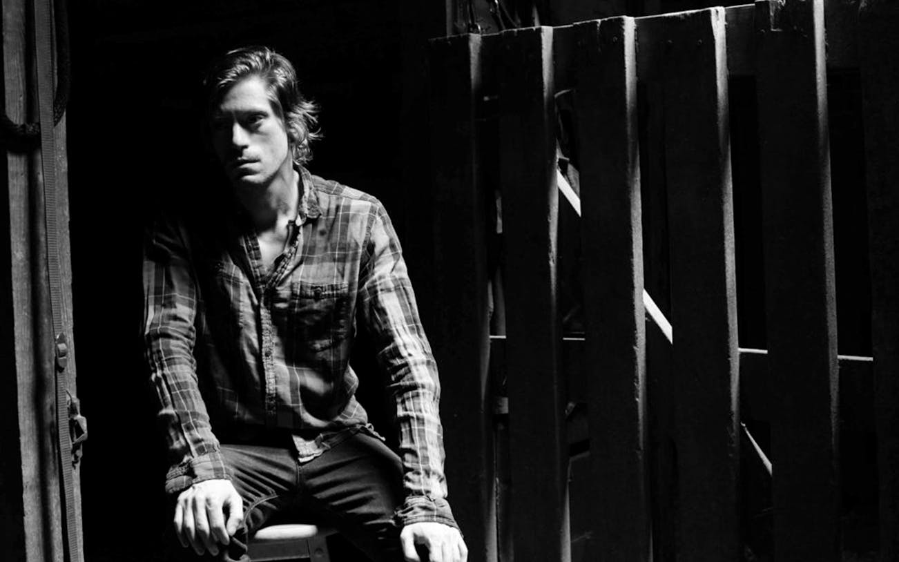 Man in plaid shirt sitting in dimly lit room, associated with Oklahoma!