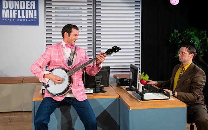 Man in pink suit plays banjo at desk in "The Office! A Musical Parody" scene.