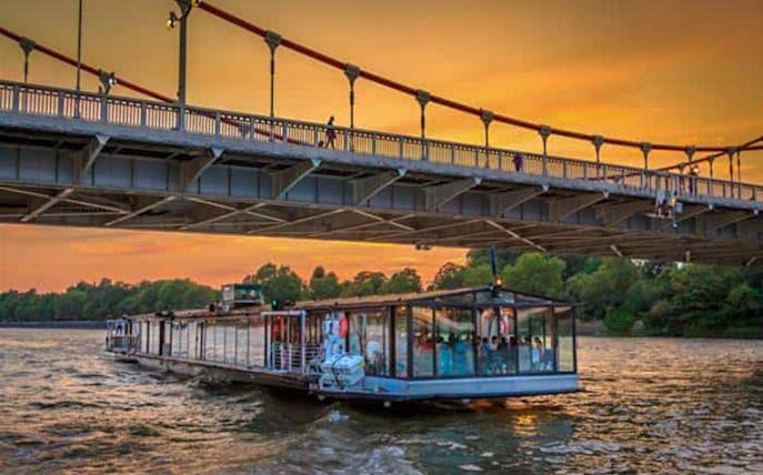 Dinner cruise boat on the Thames River in London at sunset, passing under a bridge.