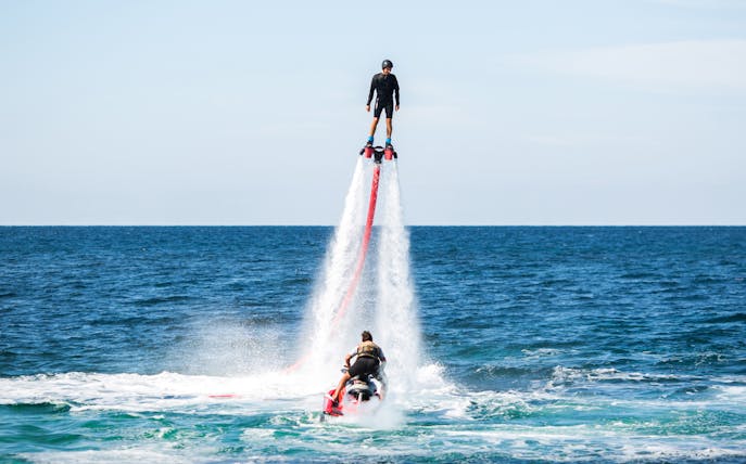Person flyboarding over ocean with jet ski in background.