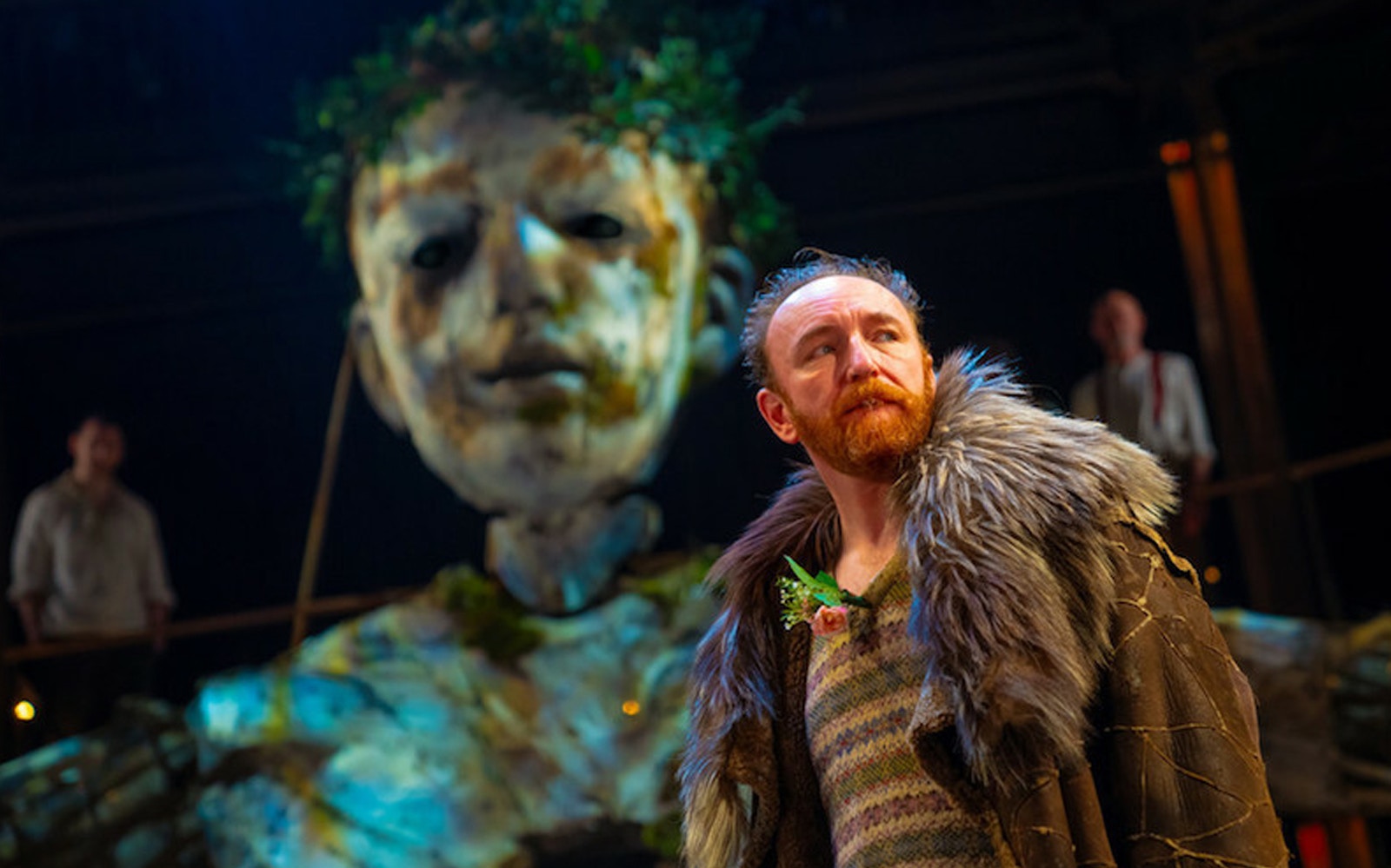 Actor in fur coat on stage with large puppet in "As You Like It" performance.