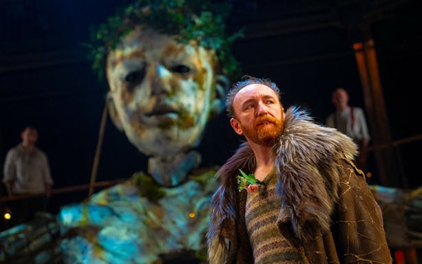 Actor in fur coat on stage with large puppet in "As You Like It" performance.