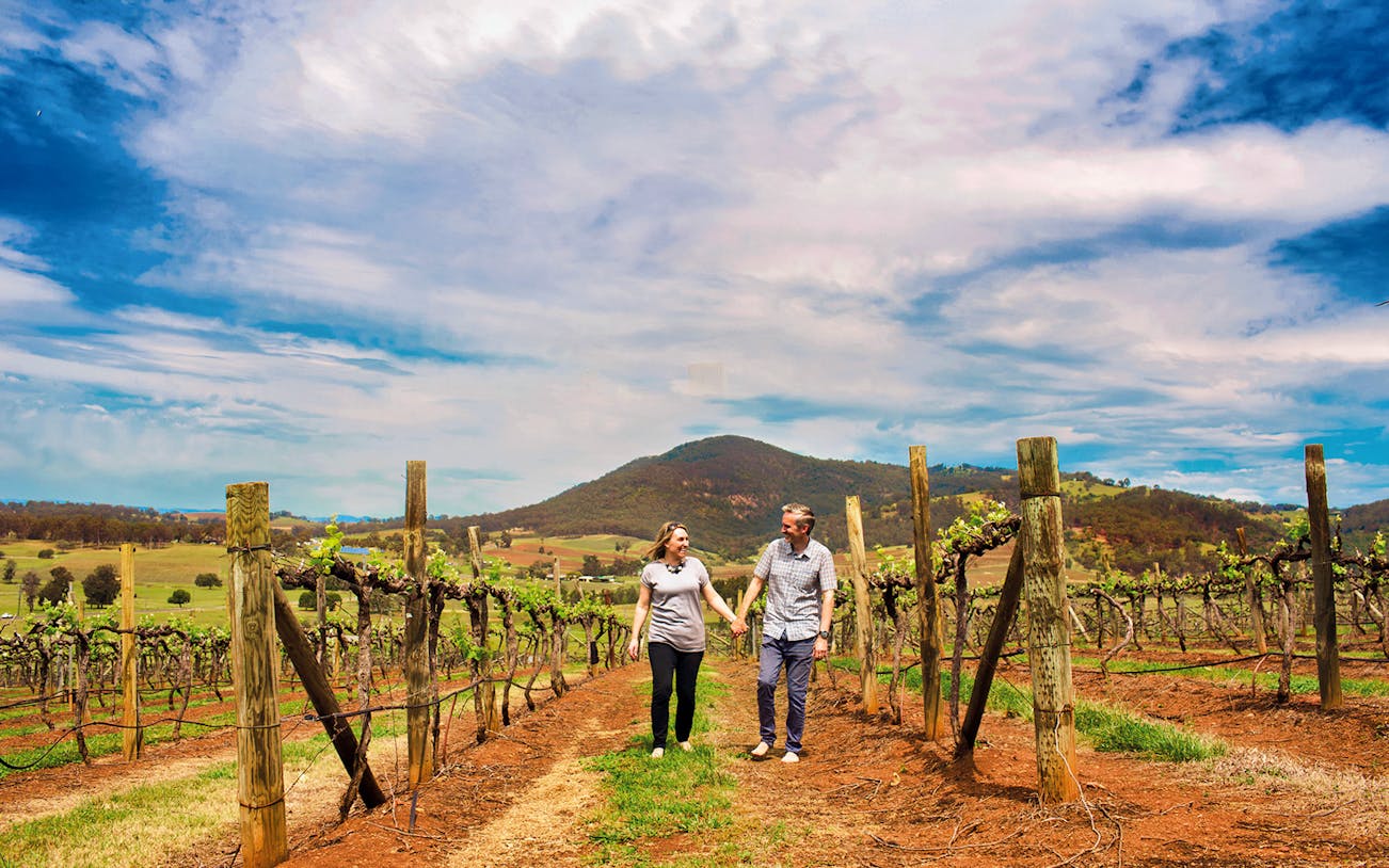 Couple walking through vineyard in Hunter Valley with scenic mountain backdrop.