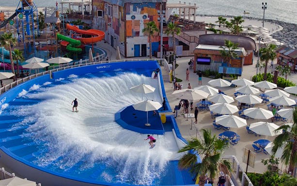 Surfers on wave simulator at Laguna Waterpark, Dubai, with slides and seating area nearby.