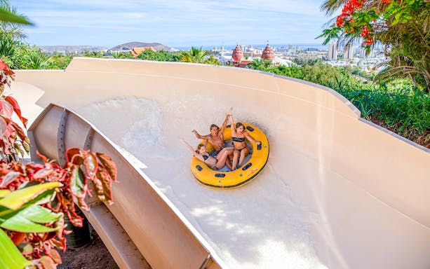Group enjoying water slide at Siam Park, Tenerife, with cityscape in background.