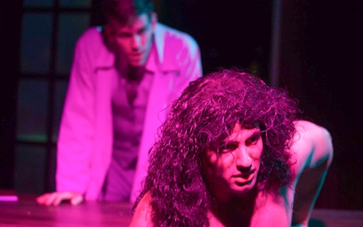 Man with long hair in dramatic lighting, another person in background, scene from Frankenstein play.