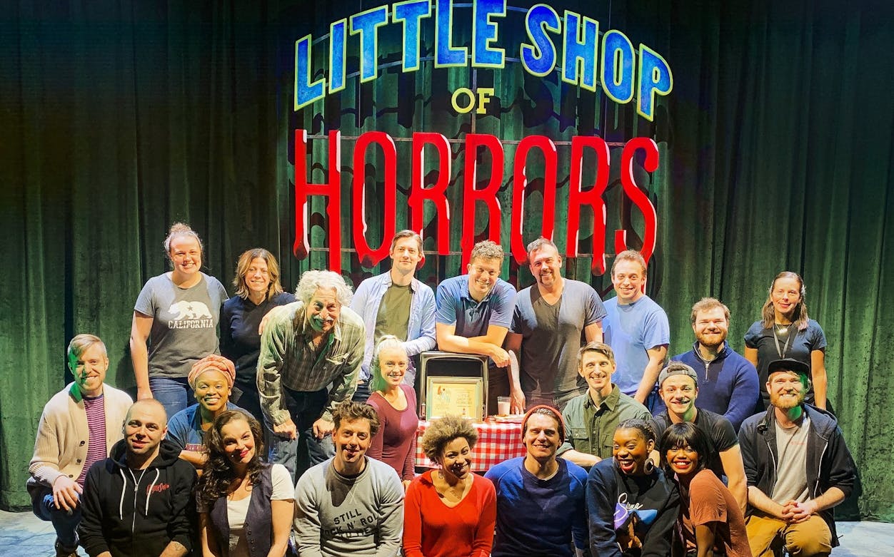 Cast of Little Shop of Horrors posing on stage with show sign in background.