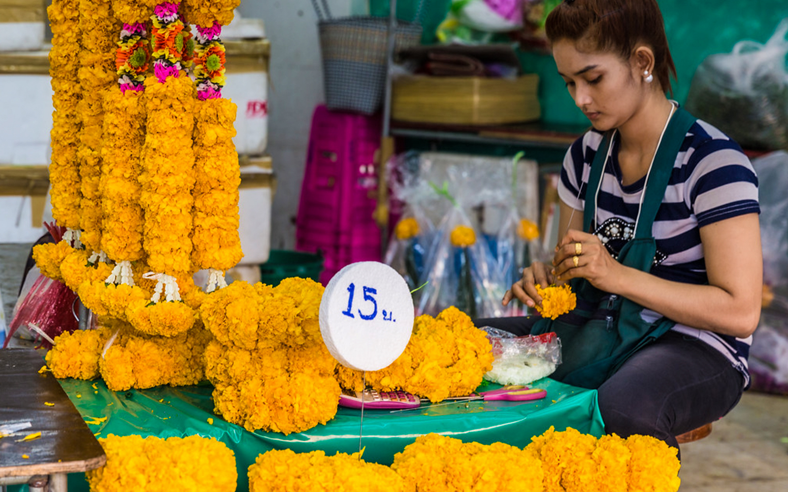 Flower garland vendor at Bangkok's Flower Market, crafting marigold garlands, price sign visible.