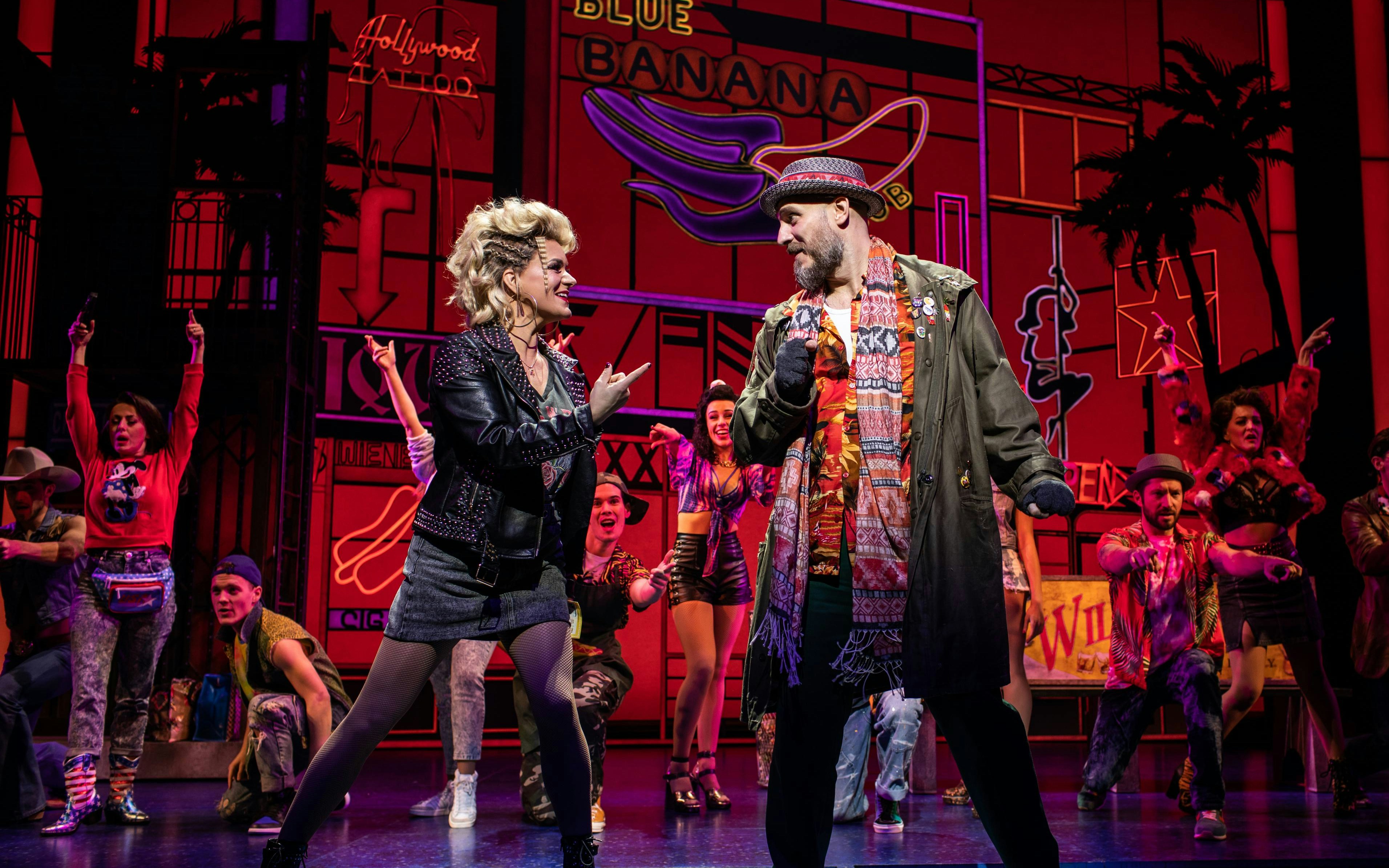 Musical cast performing on stage with neon signs in the background.