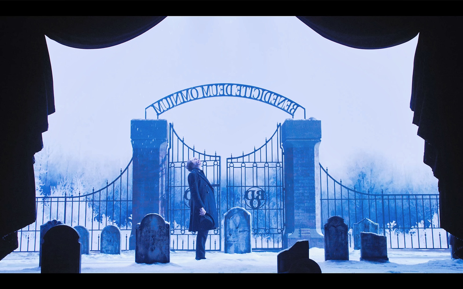 Man standing in snowy graveyard by iron gate, scene from A Christmas Carol.