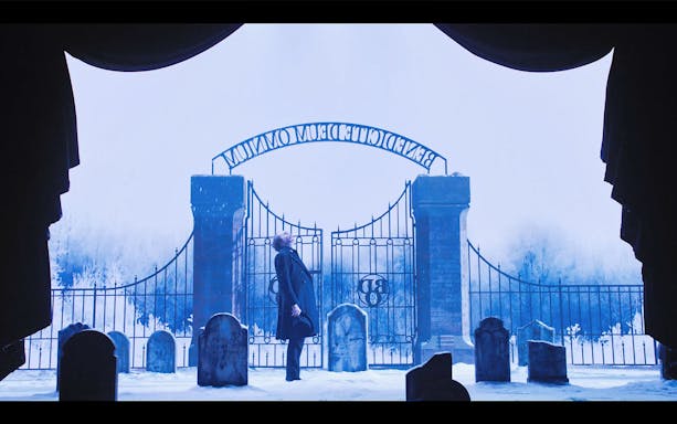 Man standing in snowy graveyard by iron gate, scene from A Christmas Carol.