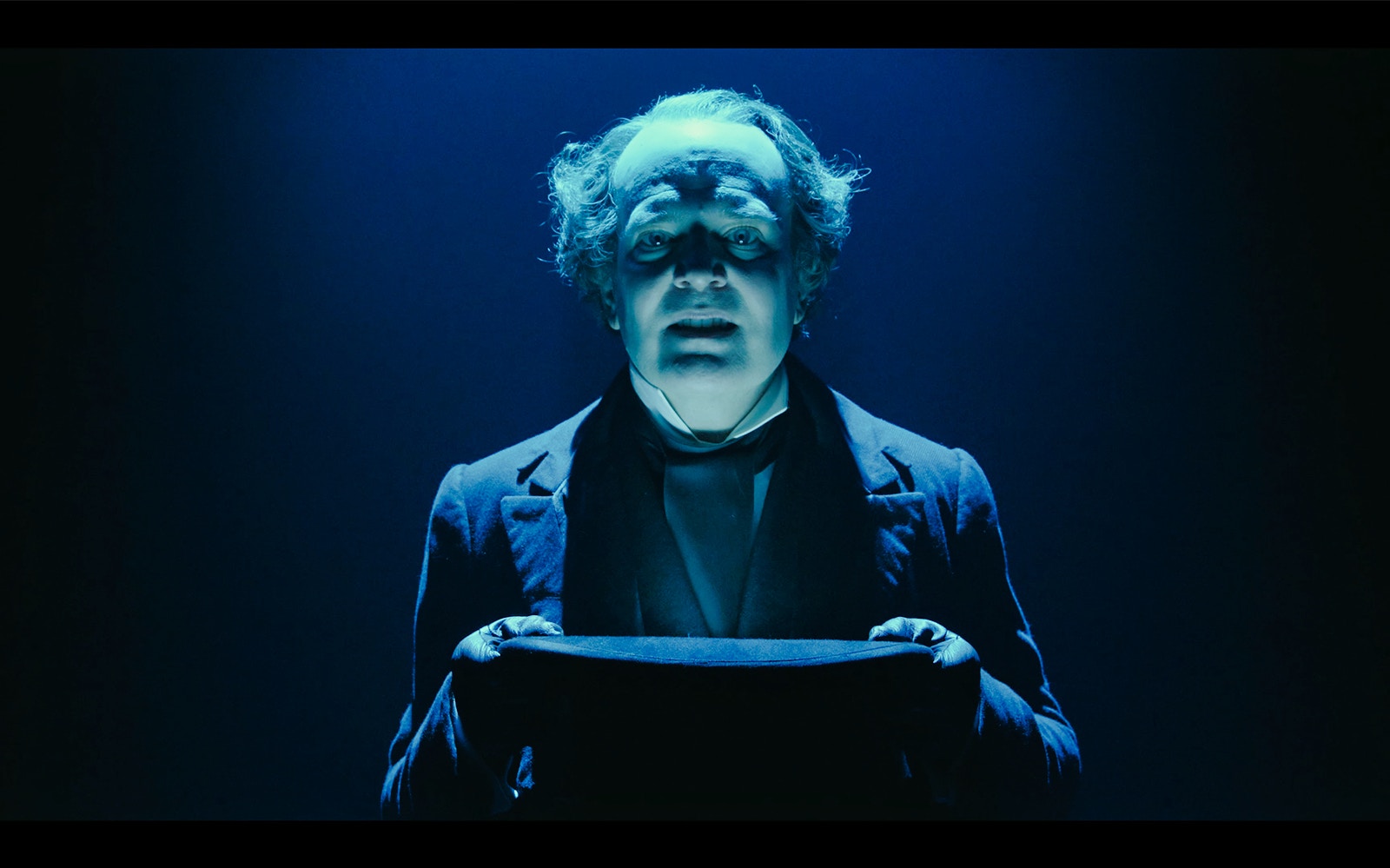 Actor in Victorian attire holding a top hat, performing in "A Christmas Carol" stage production.