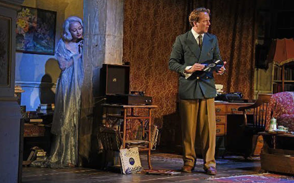Man holding a record in a vintage room, woman in a ghostly dress peeking from behind a wall, Blithe Spirit play.
